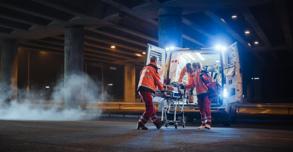 Auxiliaires medicaux chargent un patient sur civière dans une ambulance sous un pont, la nuit. Fumée au sol.