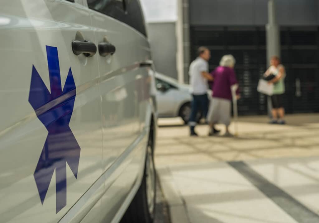 Véhicule d'urgence blanc, emblème Étoile de Vie bleue, personnes floues aidant une personne âgée avec canne.