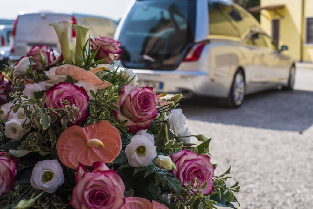 Gerbe de roses roses et blanches avec callas et feuillage, posée devant un corbillard argenté lors d'un service funéraire.